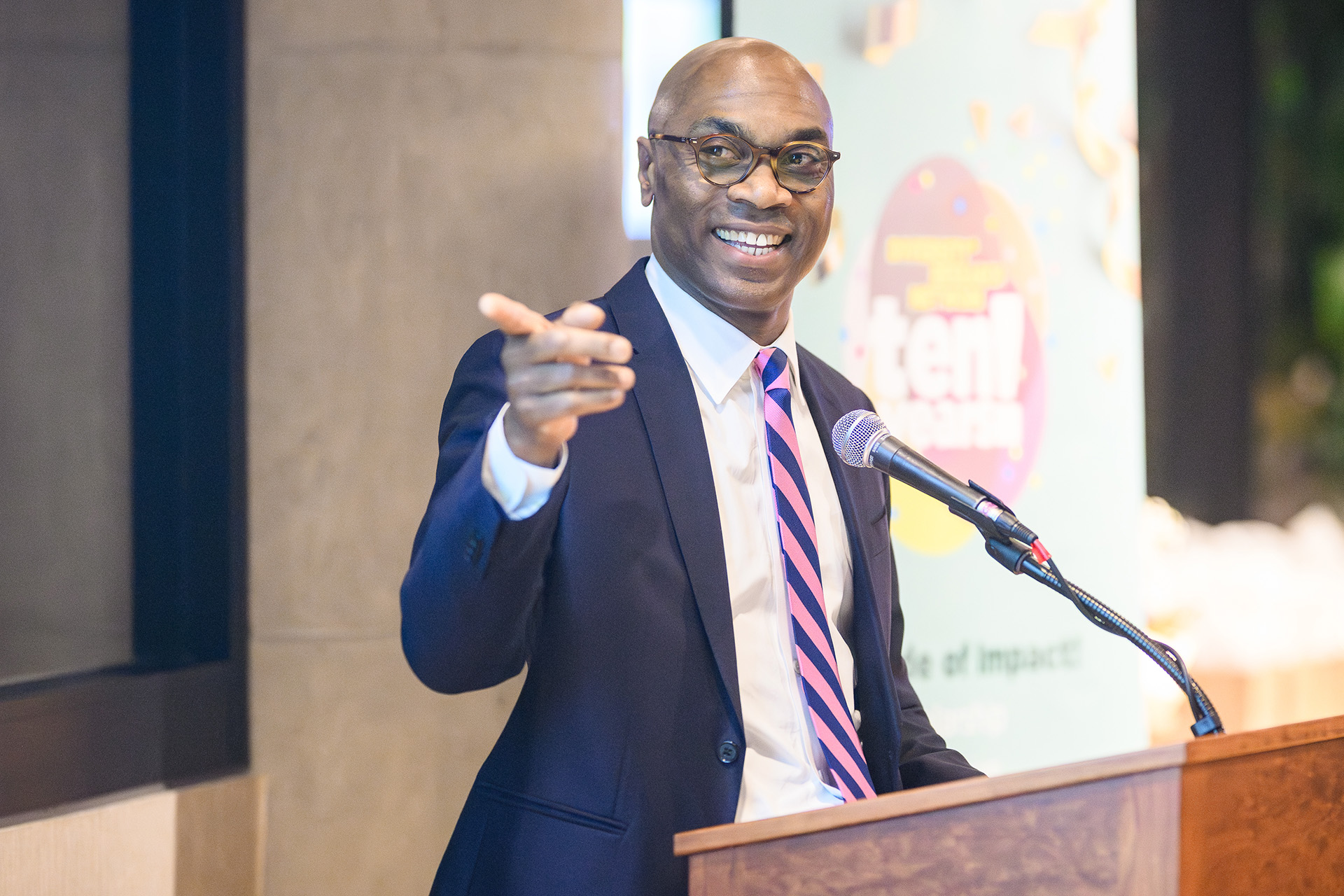 a man smiles and gestures toward the audience while speaking at the podium